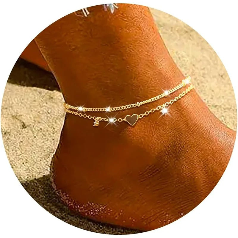 A close-up of a person's ankle wearing a delicate double-layered gold chain anklet with a small heart charm, set against a sandy beach background.