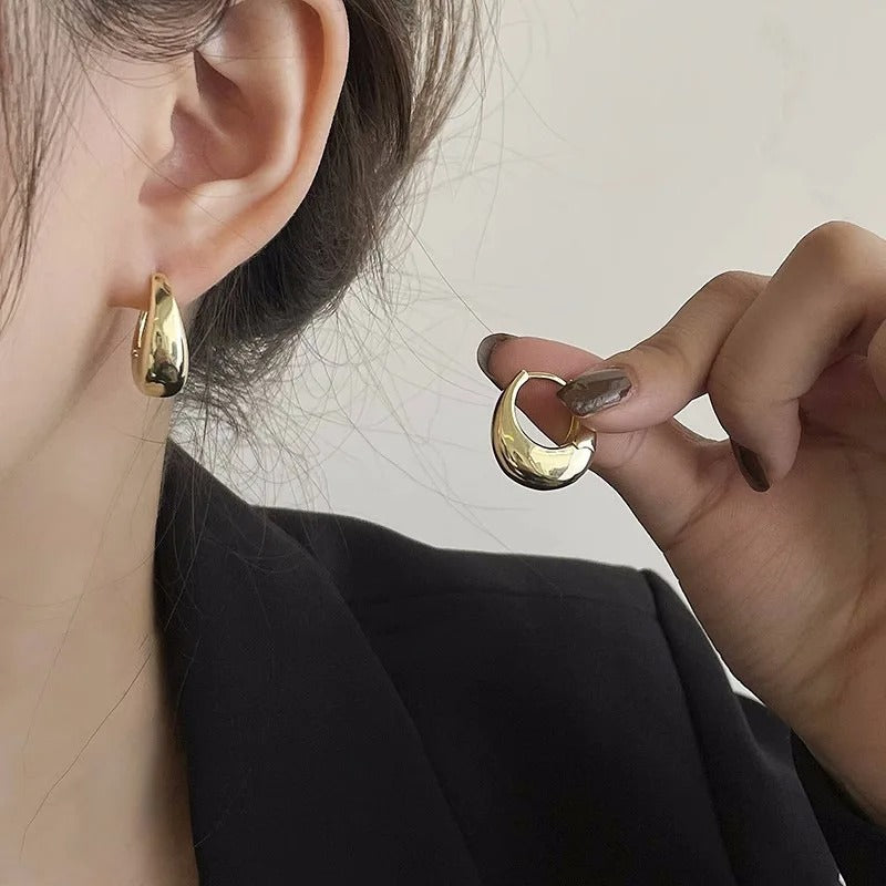 Gold hoop earrings being worn and held by a person against a neutral background