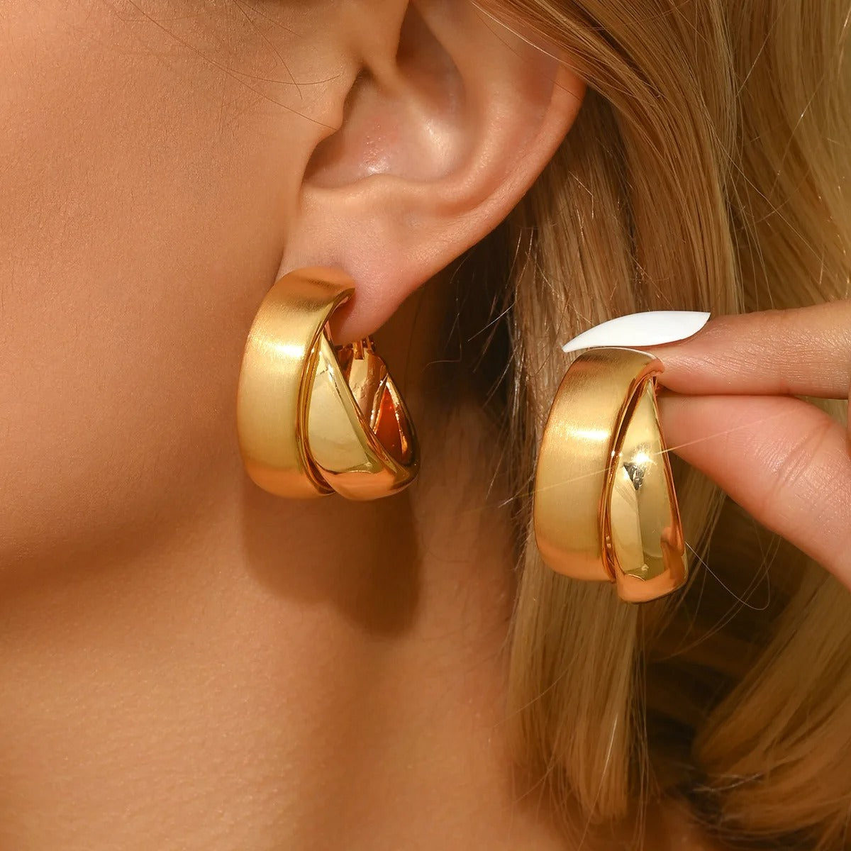 A close-up of a person wearing and holding a pair of polished gold hoop earrings. The earrings have a thick, rounded design with a reflective finish, shown on the ear and held by fingers with a white manicured nail.