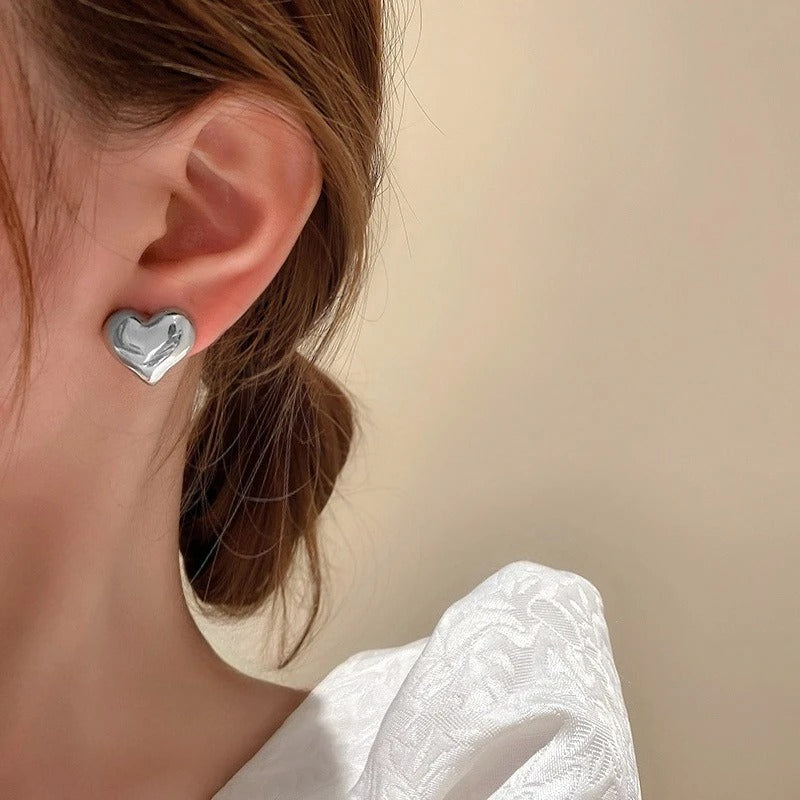 A close-up of a person's ear wearing a small silver heart-shaped stud earring, with brown hair tied back and a white textured top against a plain beige background.