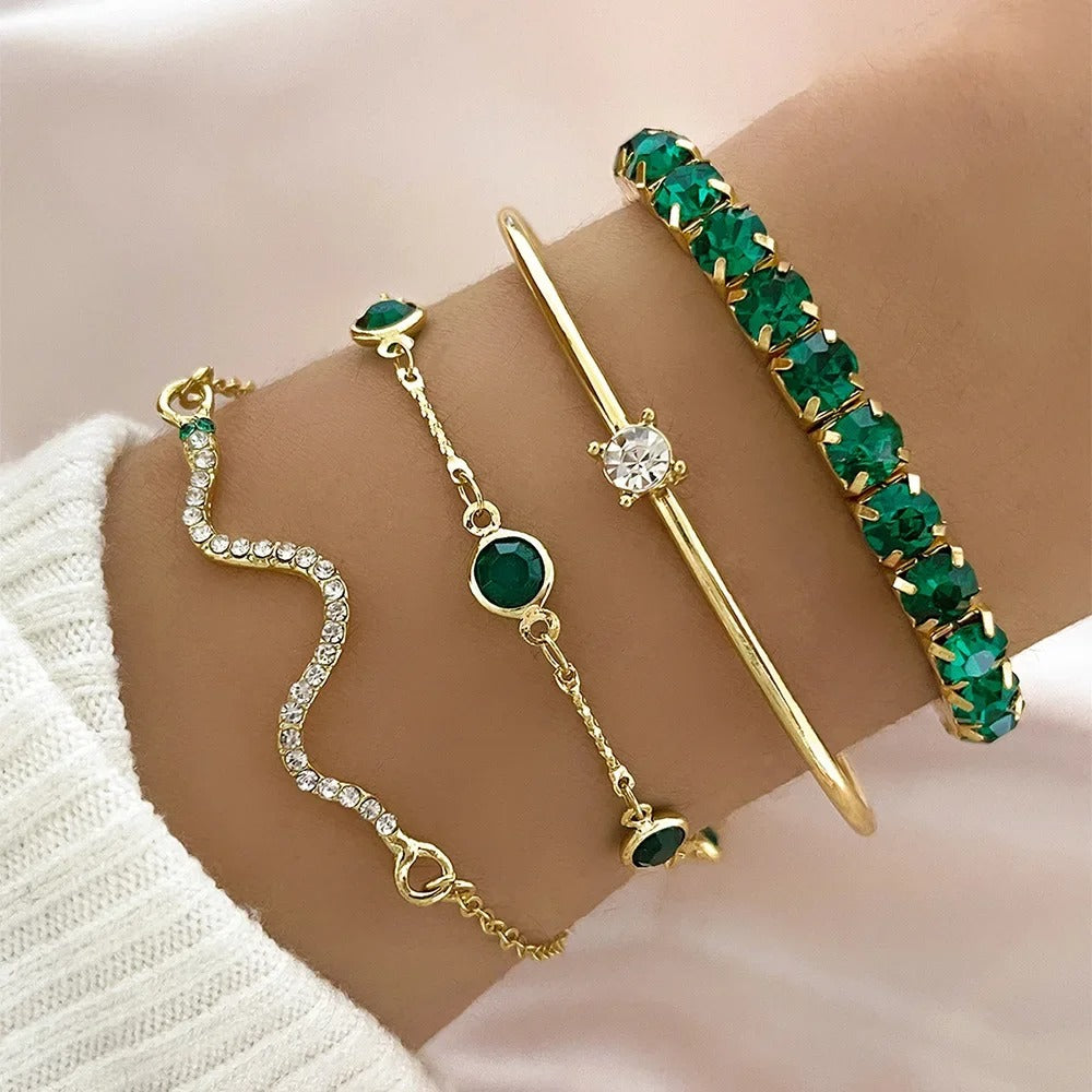 Four gold-toned bracelets displayed on a person's wrist, including a wavy crystal bracelet, a chain bracelet with green stones, a slim bar bracelet with a clear center stone, and a bangle covered in green stones.
