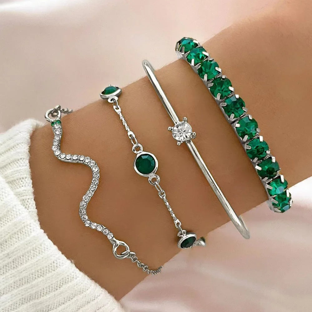 Four gold-toned bracelets displayed on a person's wrist, including a wavy crystal bracelet, a chain bracelet with green stones, a slim bar bracelet with a clear center stone, and a bangle covered in green stones.