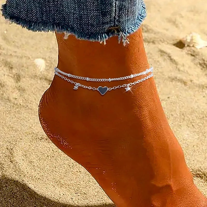 A close-up of a person's ankle and foot on a sandy beach, wearing a delicate silver chain anklet with a small blue heart charm.