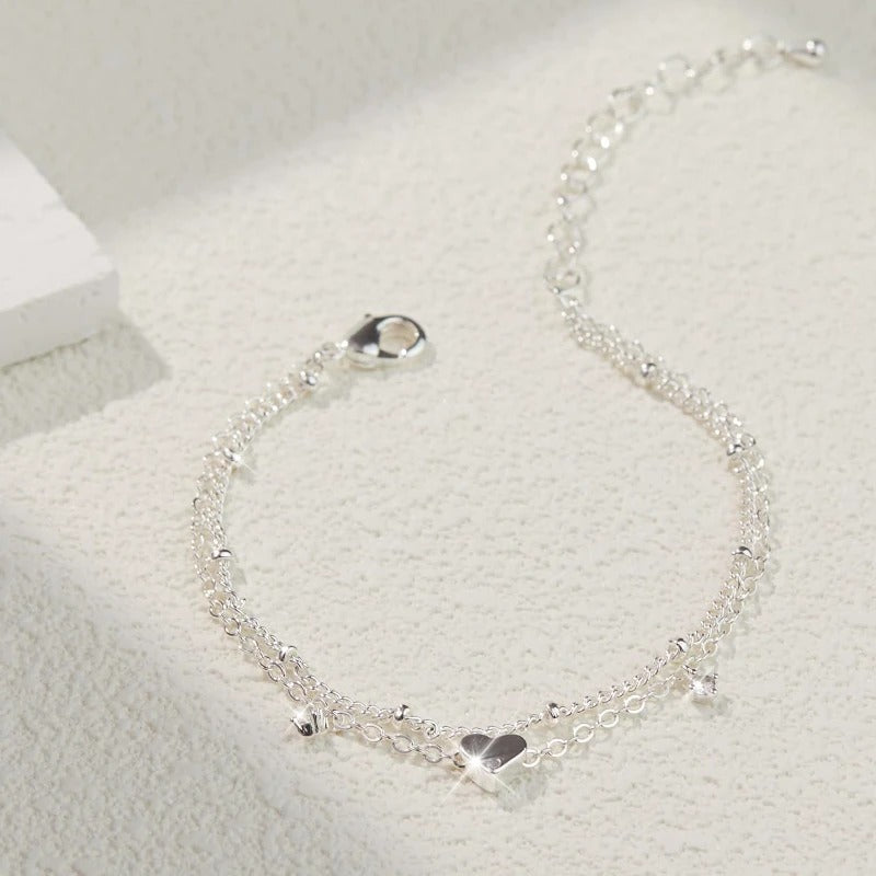 A delicate silver chain anklet with a small heart charm and tiny dangling beads, displayed on a textured white surface.