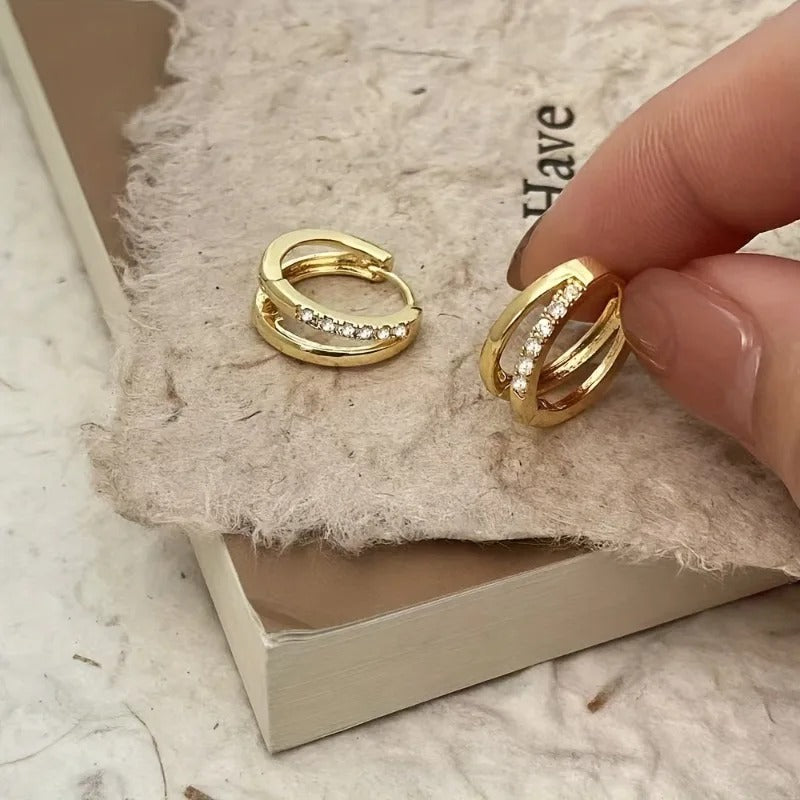 Gold hoop earrings with embedded stones held by a hand on a textured surface.