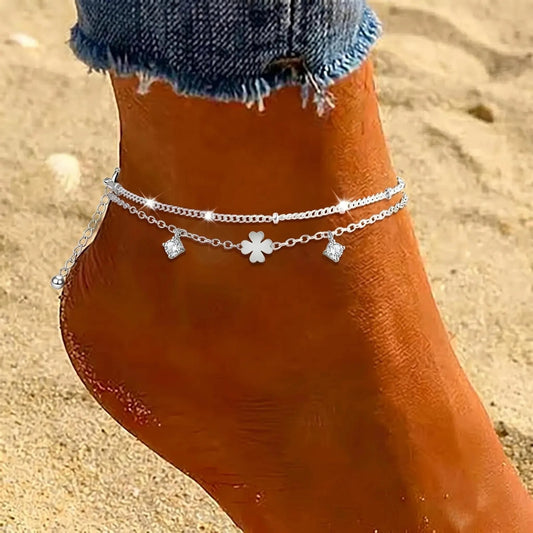 A close-up of a person's ankle and foot wearing a delicate double-strand silver anklet with a central flower charm and small dangling charms, photographed outdoors on sand with denim-clad lower legs visible.