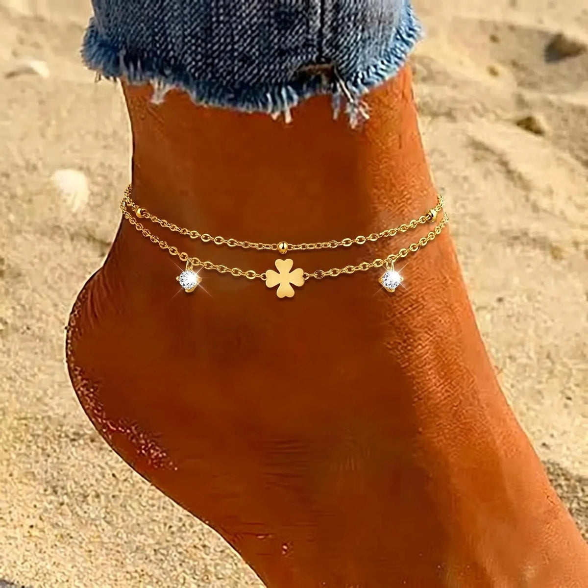 A close-up of a person's ankle and foot wearing a delicate double-layered gold anklet with a central four-leaf clover charm and two small sparkling rhinestone accents, set against a sandy beach background.